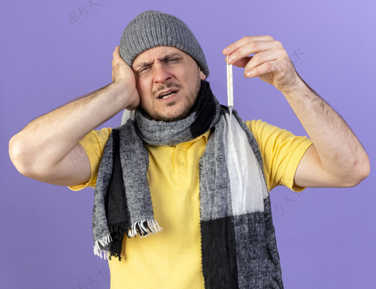 斯拉夫人一个戴着冬天帽子和围巾的年轻金发病患把手放在头上 把温度计隔离在紫色的墙上疾病围巾姿势