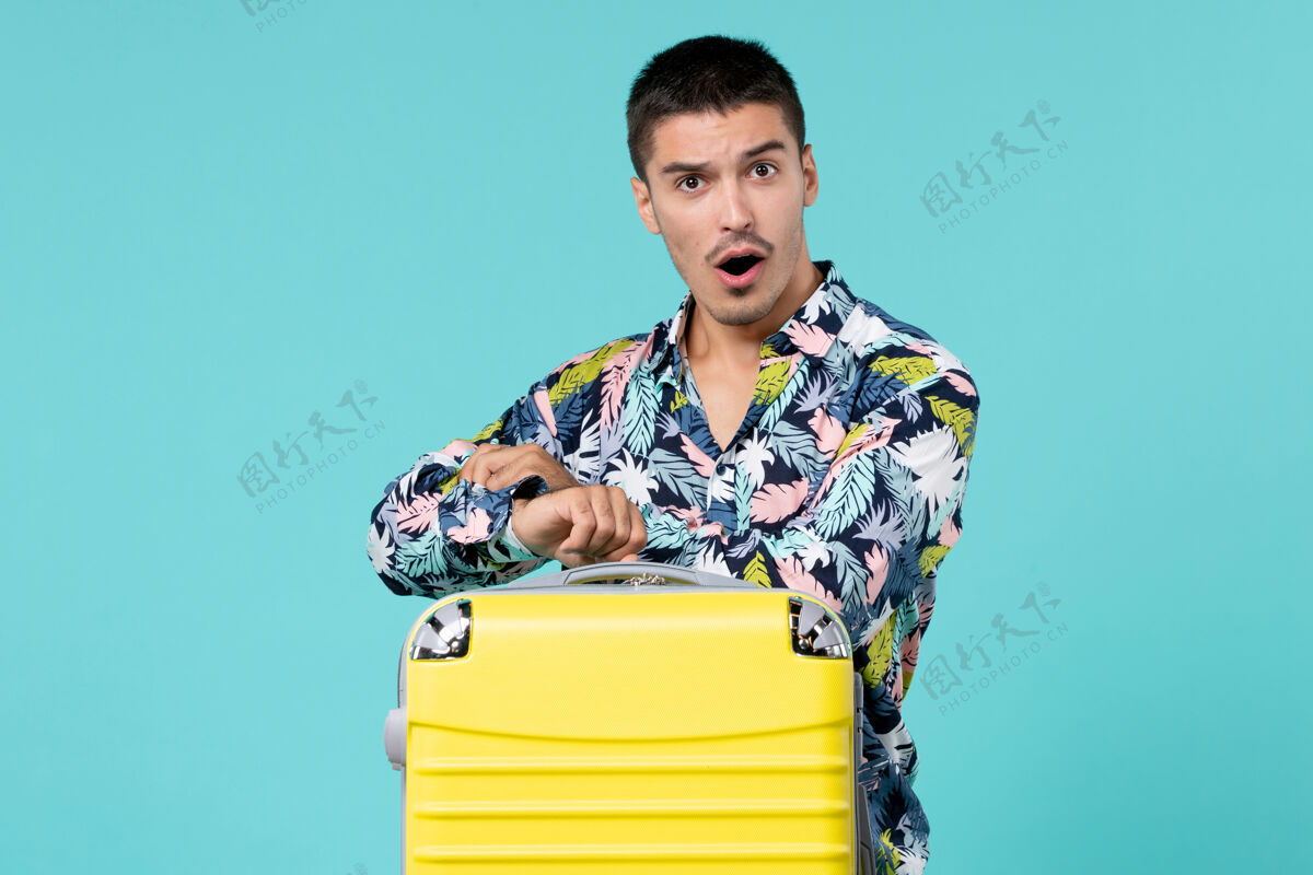 旅程年轻的男性正拿着他的黄色袋子准备旅行 在蓝色的墙上查看时间航行微笑人