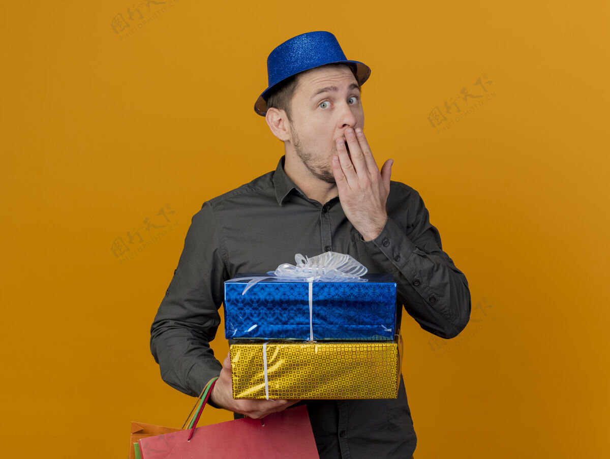 年轻惊讶的年轻人戴着蓝色的帽子拿着礼品盒和袋子 用手捂住嘴巴孤立在橙色派对惊喜蓝色