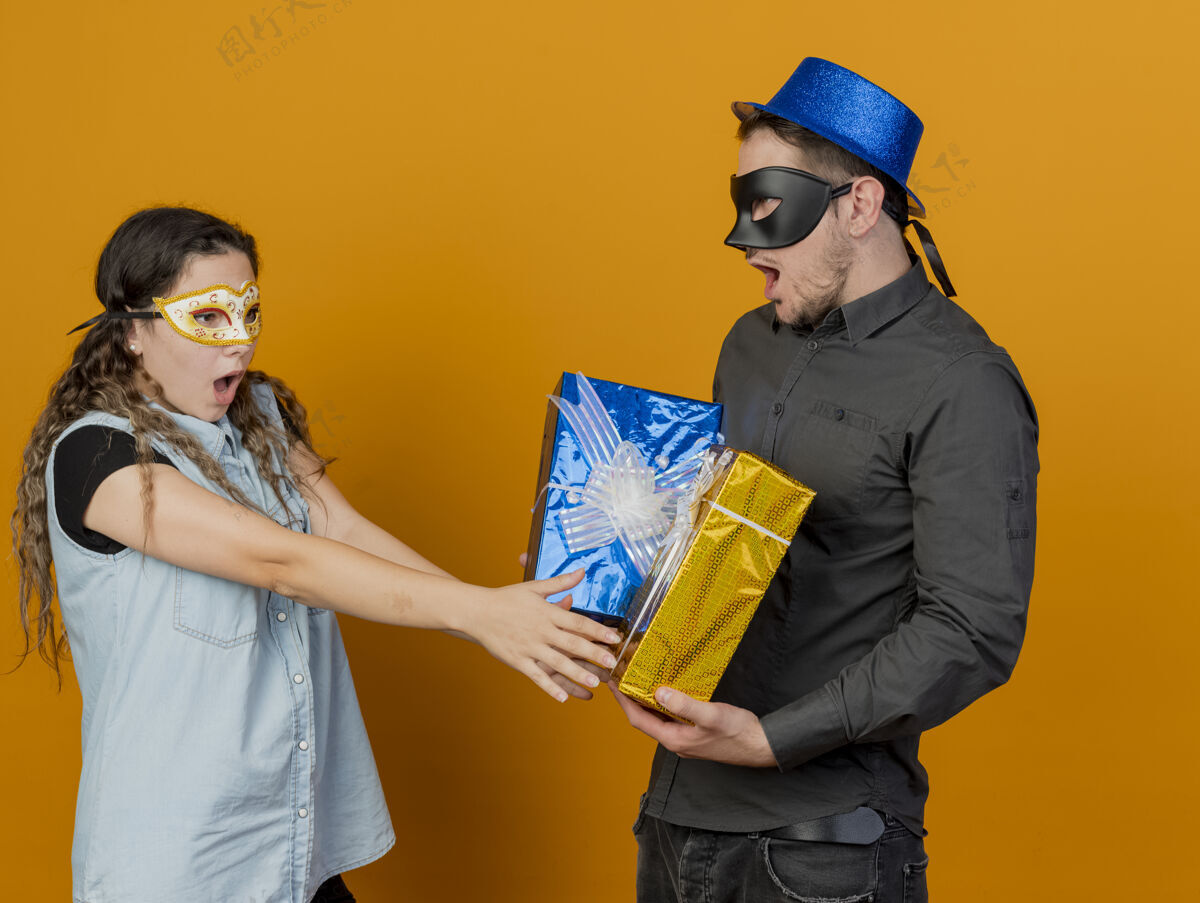 穿惊讶的年轻人戴着化装舞会眼罩女孩伸出手在礼物在他的手上孤立的橙色化装舞会男人眼睛