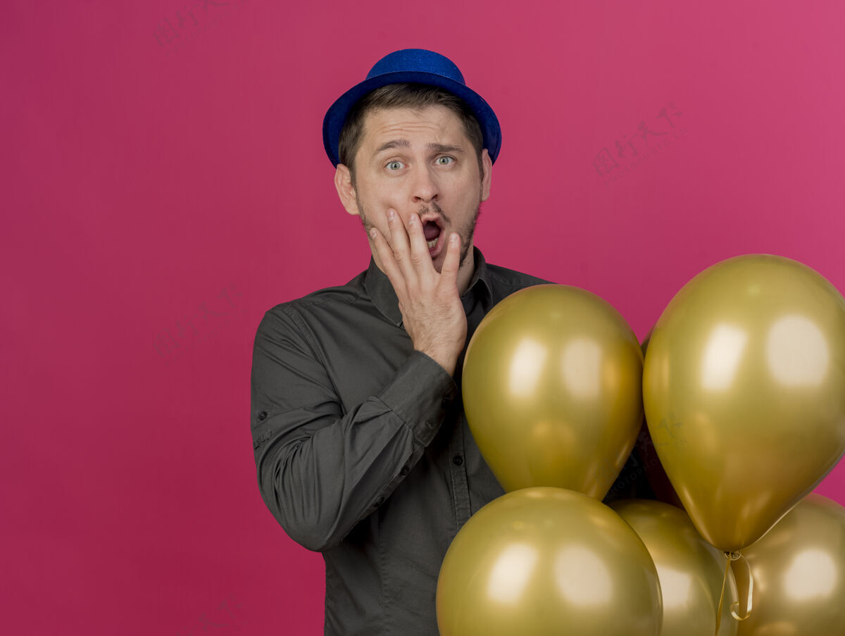 嘴巴一个戴着蓝色帽子的年轻人站在气球旁边 手被粉色隔离 嘴巴被气球盖住了小伙子派对蓝色