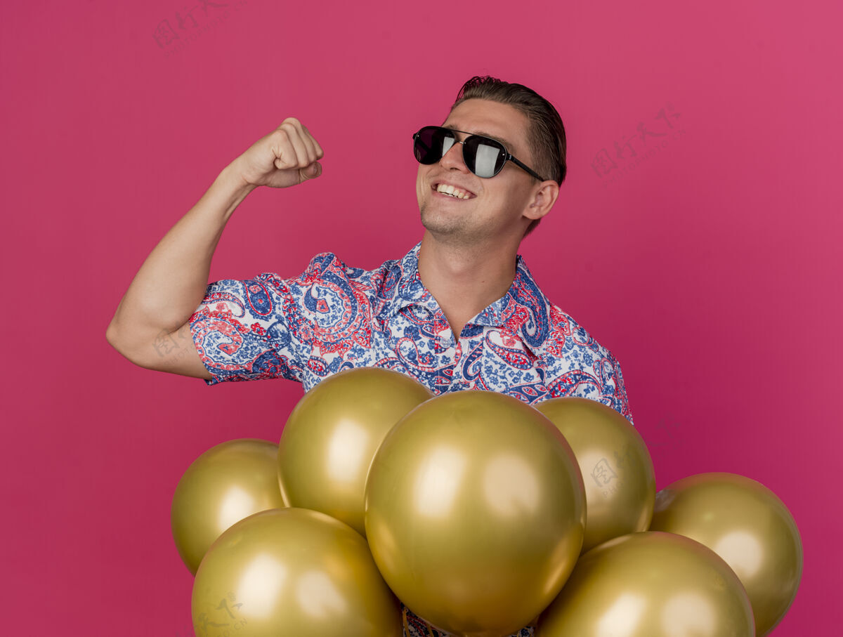 侧线微笑着的年轻人穿着五颜六色的衬衫站在气球后面 在粉红色的地板上做着“是”的手势气球站粉色