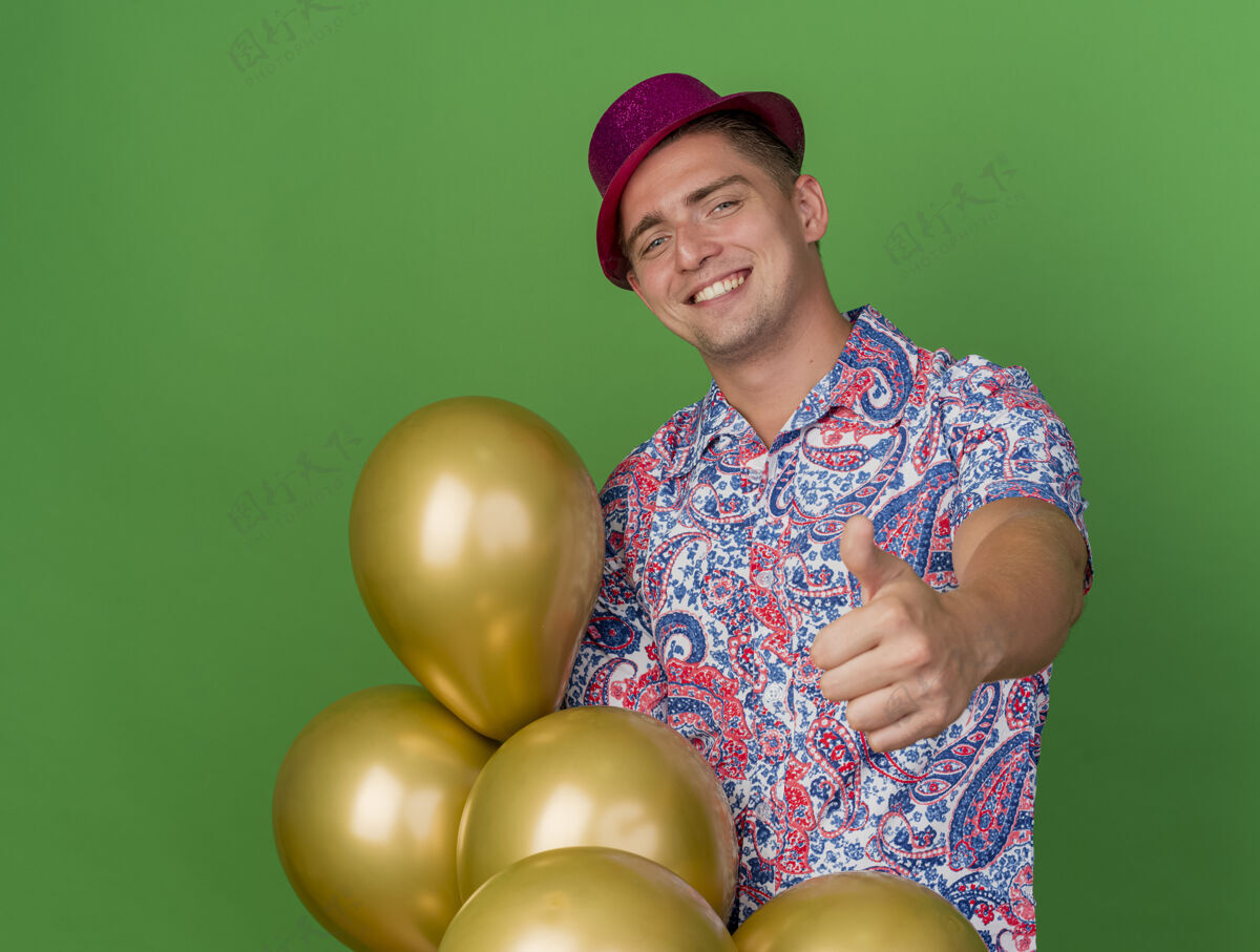绿色微笑着的年轻人戴着粉红色的帽子 拿着气球 在绿色的草地上孤立地竖起大拇指粉色帽子穿