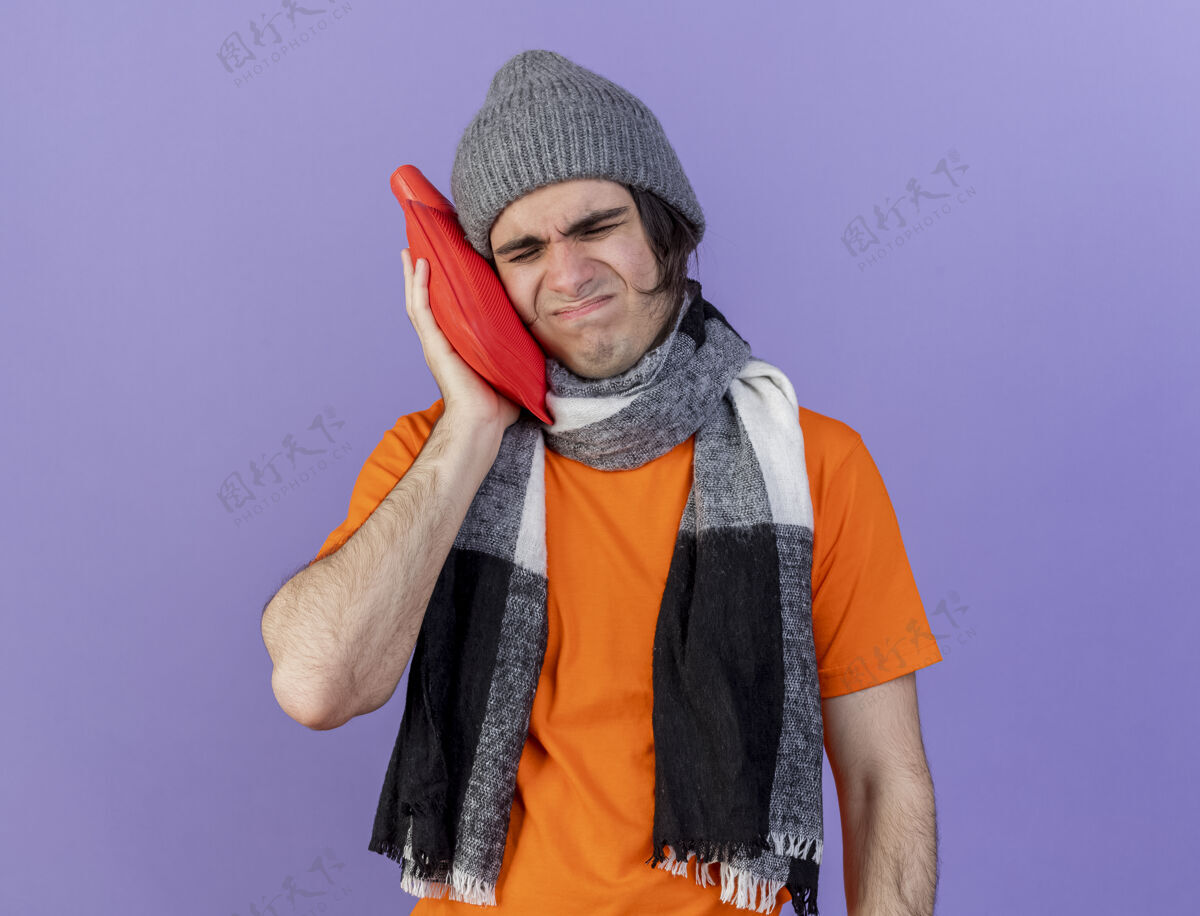 围巾未出狱的年轻病夫戴着冬天的帽子戴着围巾把热水袋放在脸颊上隔离在紫色上男人不愉快水