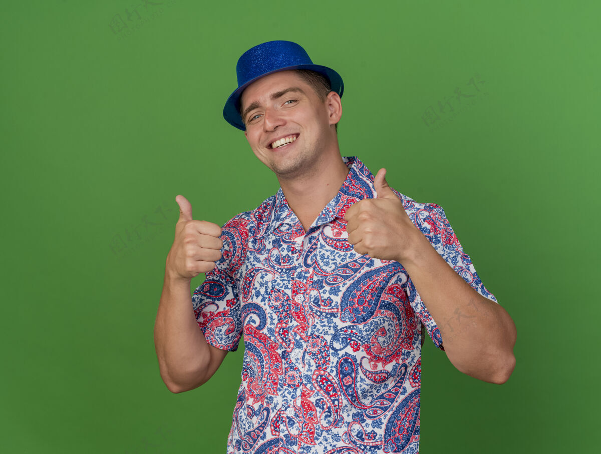 派对戴着蓝色帽子的年轻人微笑着 在绿色的草坪上竖起大拇指穿帽子拇指