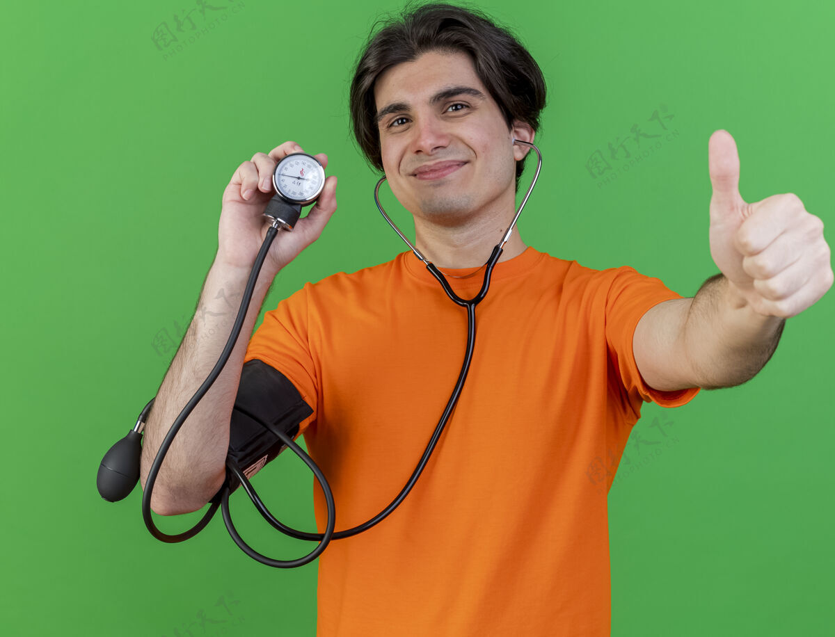 显示高兴的年轻人戴着听诊器拿着血压计 在绿色的草坪上孤立地竖起大拇指年轻绿色疾病