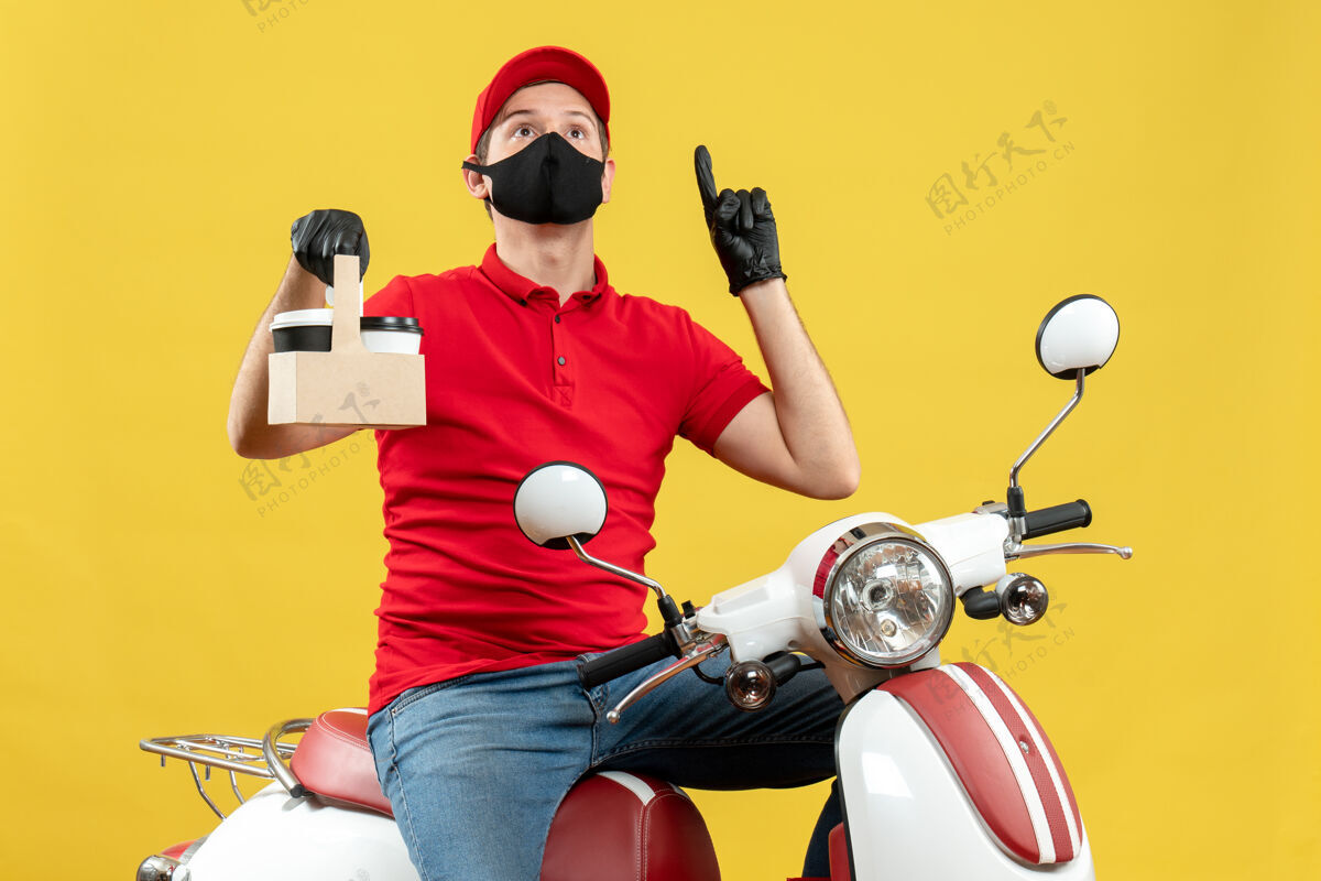 命令俯视图：身穿红色上衣 戴着帽子手套 戴着医用面罩的快递员坐在滑板车上 手里拿着指向上的订单摩托车手套男子