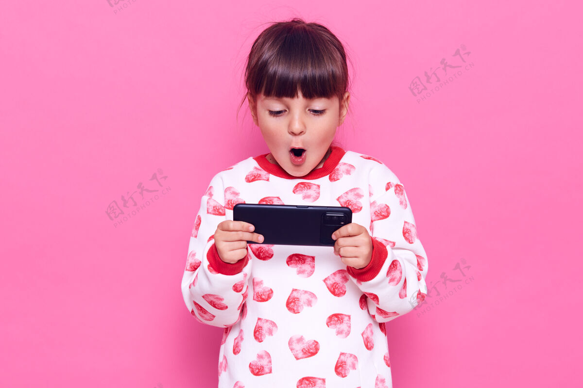 惊喜一头黑发的迷人小女孩 一头黑发 张开嘴拿着智能手机 玩游戏 对结果感到惊讶 在粉红色的墙上摆出孤立的姿势人信息兴奋