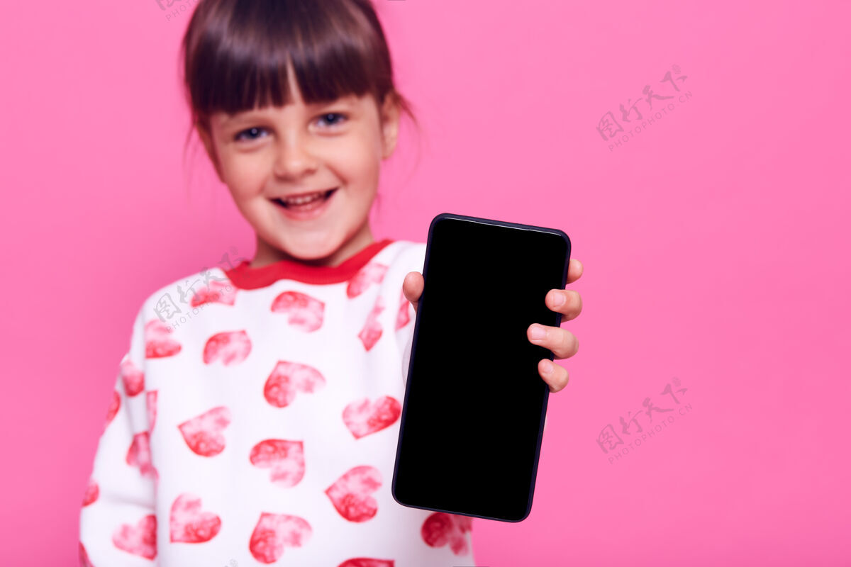 打字可爱的快乐微笑的小女孩穿着休闲风格的套头衫 手里拿着手机的空白屏幕 在粉色的墙上摆出孤立的姿势愉快可爱孩子