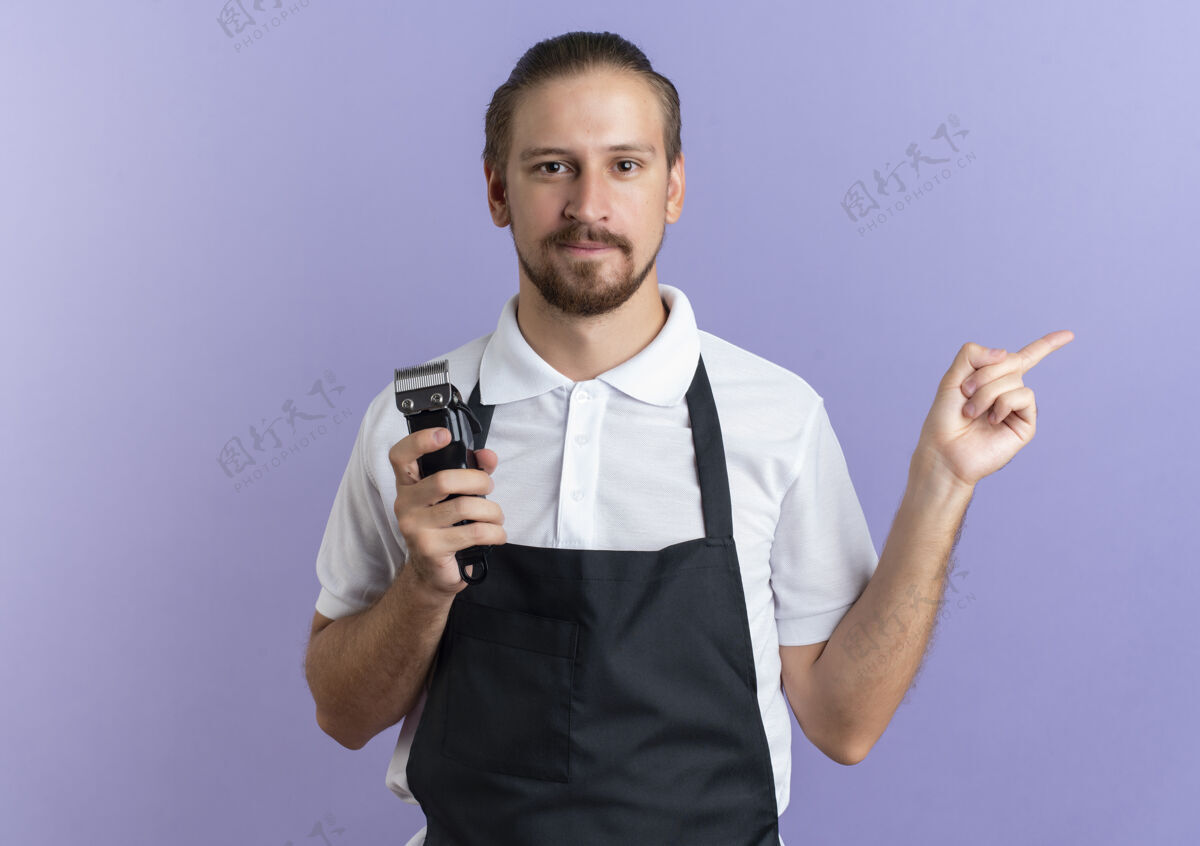 拿着年轻帅气的理发师穿着制服 手里拿着理发器 指着隔离在紫色墙上的一边紫色制服剪子