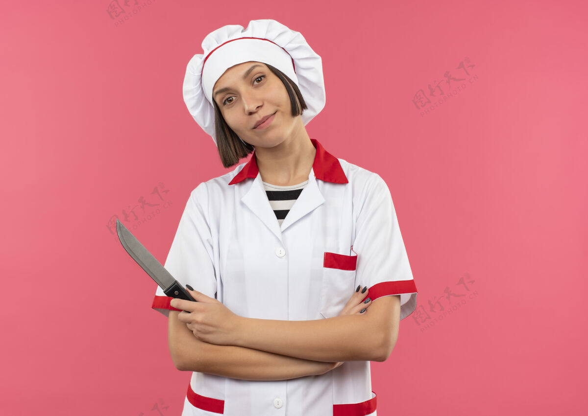 拿着身着厨师制服的年轻女厨师站在粉红色的墙上 手拿刀 姿势封闭闭合衣服请
