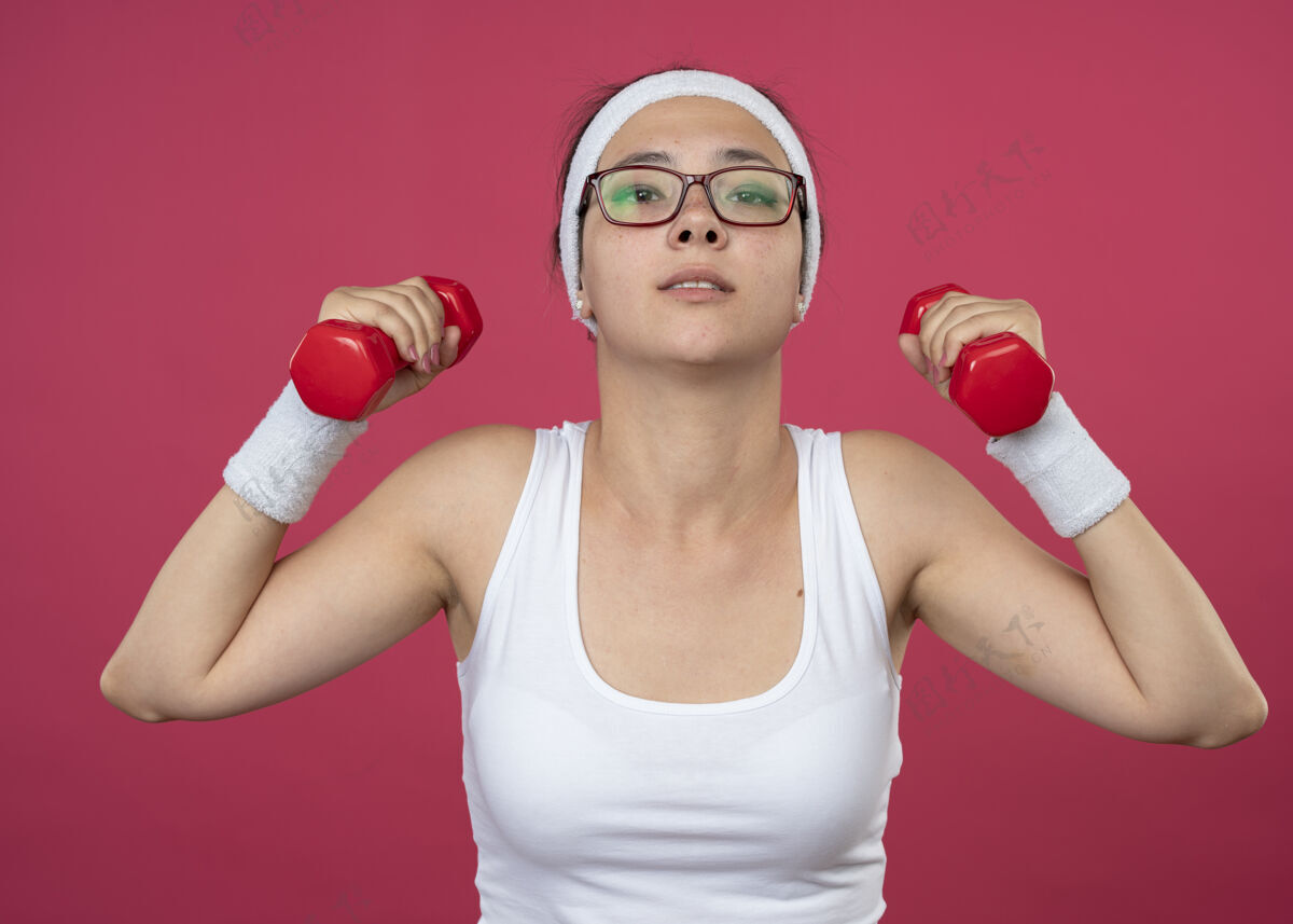 自信戴着眼镜 戴着头带和腕带的自信的年轻运动女性 在粉红色的墙上孤立地举着哑铃戴眼镜人