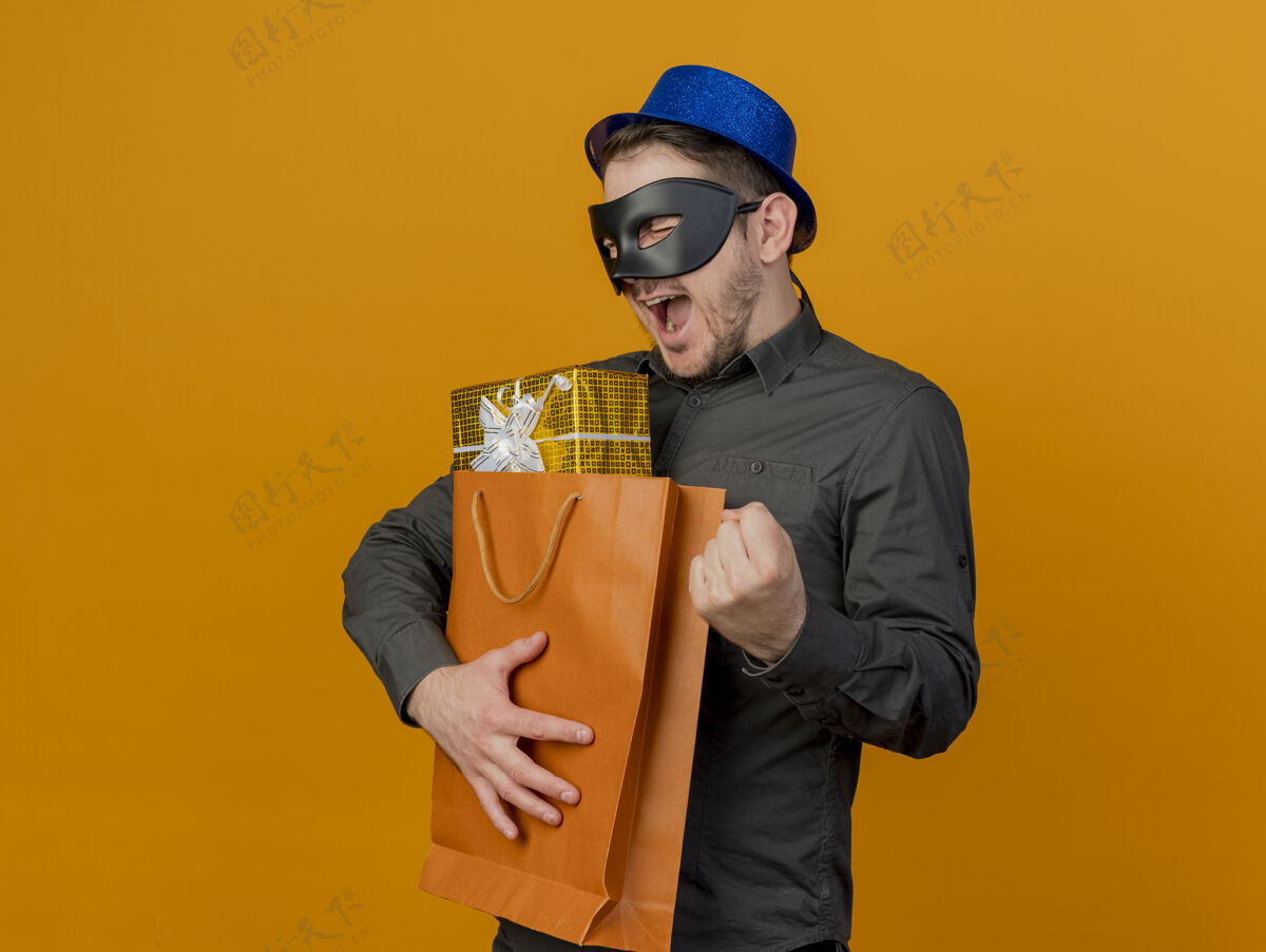 眼睛快乐的年轻人戴着蓝色帽子 戴着化装眼罩 拿着礼品袋 在橙色上显示“是”的手势年轻人穿橙色