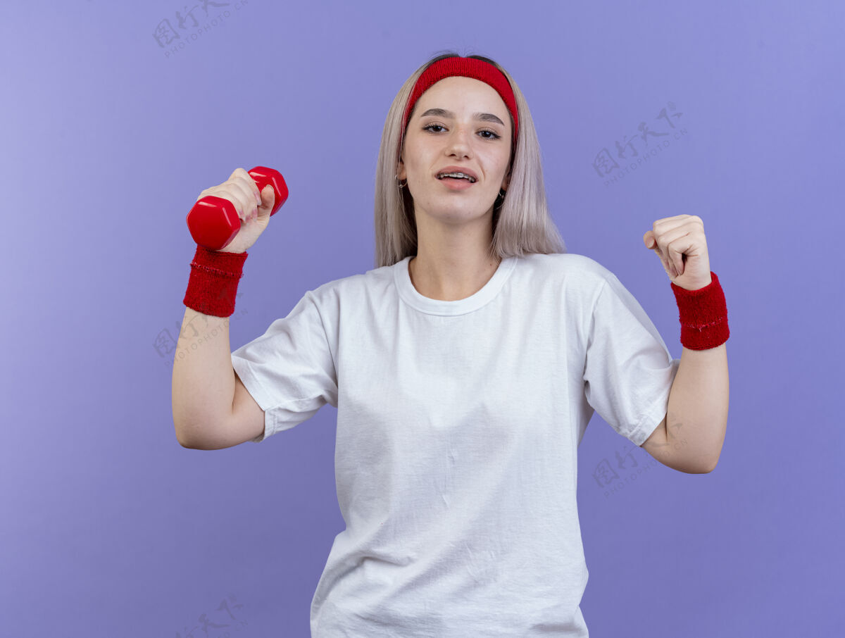 人戴着头带和腕带的戴着背带的快乐的年轻运动女性保持着拳头 在紫色的墙上孤立地举着哑铃请腕带哑铃