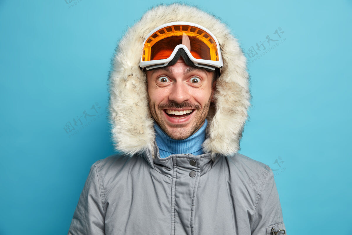 护目镜开朗的未刮胡子的男人 喜出望外 笑容满面 戴着滑雪镜 带兜帽的冬季夹克 享受极限冬季运动夹克极限休闲