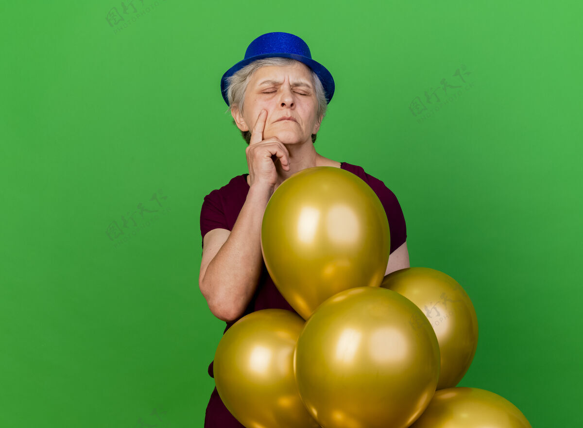 老人戴着派对帽的老太太手放在下巴上站着 氦气球被隔离在绿色墙壁上 留有复印空间下巴穿女人