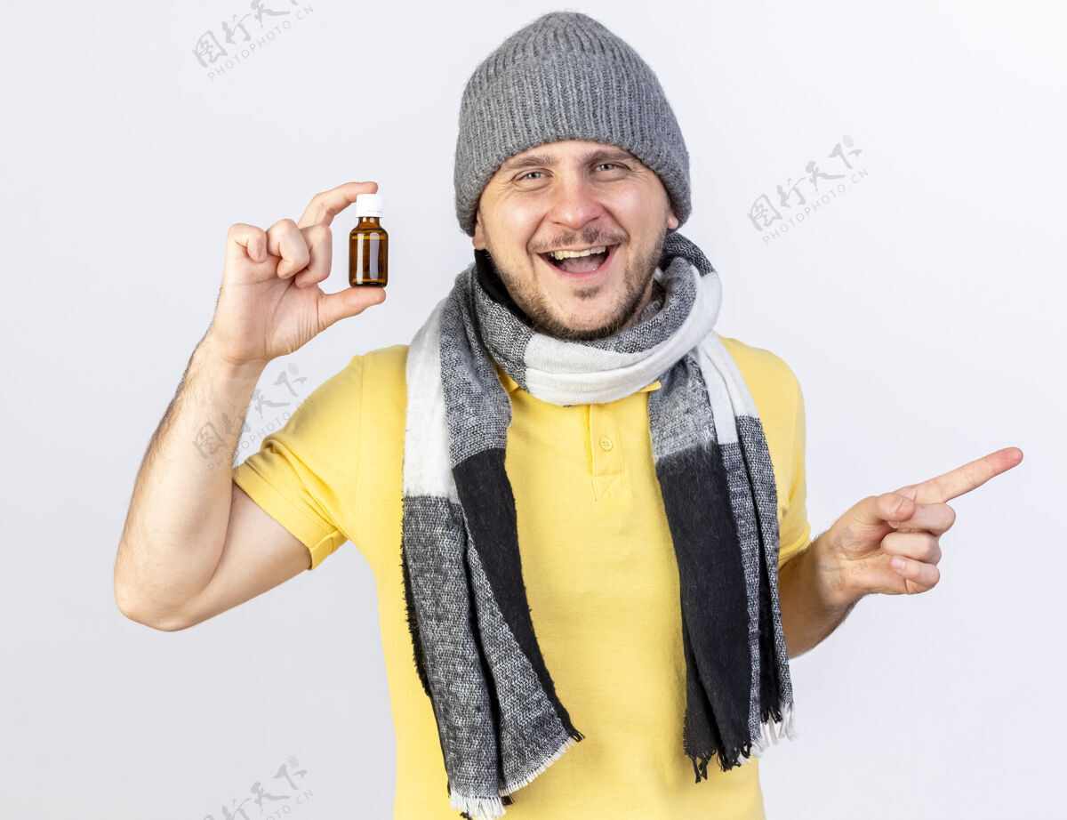 瓶子快乐的年轻金发病斯拉夫男子戴着冬季帽子和围巾拿着在玻璃瓶中的药指着隔离在白色墙壁与复制空间的一面指尖举行疾病