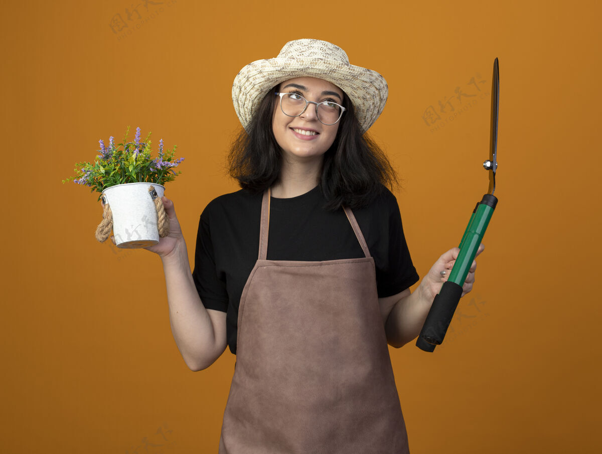 花园带着微笑的年轻黑发女园丁 戴着眼镜 穿着制服 戴着园艺帽 把花园剪子和花盆孤立地放在橙色的墙上 留着复制空间制服深色穿