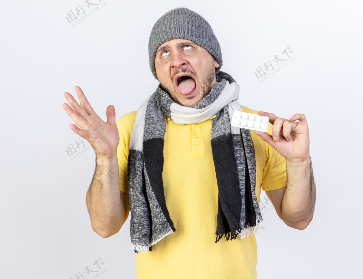 金发女郎恼怒的年轻金发病斯拉夫男子戴着冬天的帽子和围巾站起来 举起手拿着一包药丸 看着隔离在白色墙上的复制空间站着围巾帽子