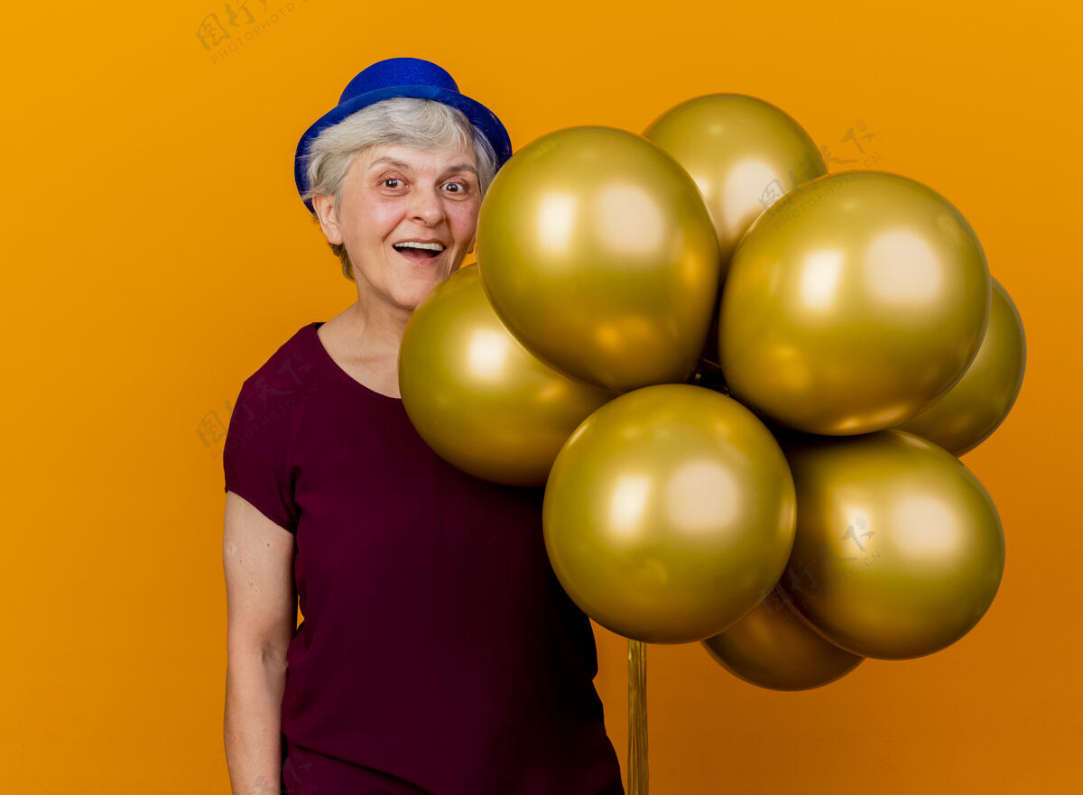 女人快乐的老妇人戴着聚会帽 手里拿着氦气球 孤立地放在橙色的墙上 还有复制空间拿穿气球