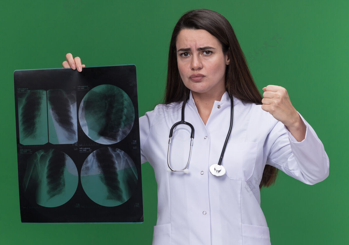 年轻愤怒的年轻女医生穿着医用长袍 手持听诊器 握紧拳头 把x光片结果放在绿色的纸上医生拳头女性