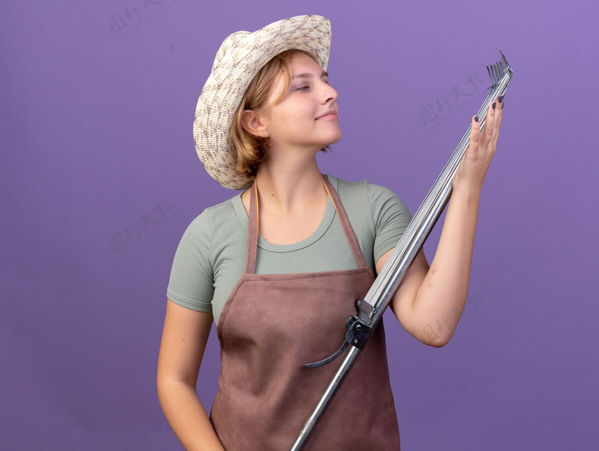 拜托高兴的年轻斯拉夫女园丁戴着园艺帽 拿着紫色的耙子看着树叶女的斯拉夫人帽子