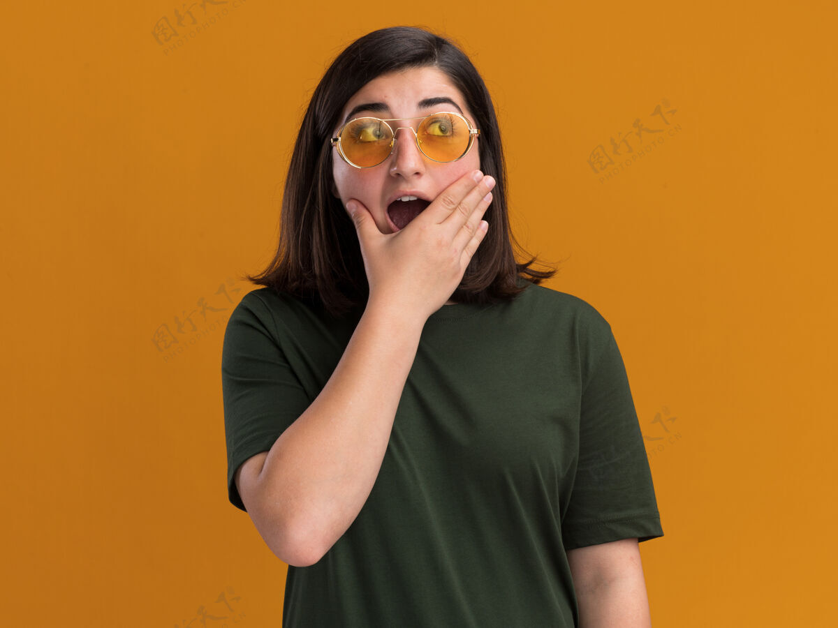 手一个戴着太阳眼镜的年轻漂亮的白人女孩惊讶地把手放在嘴巴上 看着橙色的一边年轻眼镜橙色
