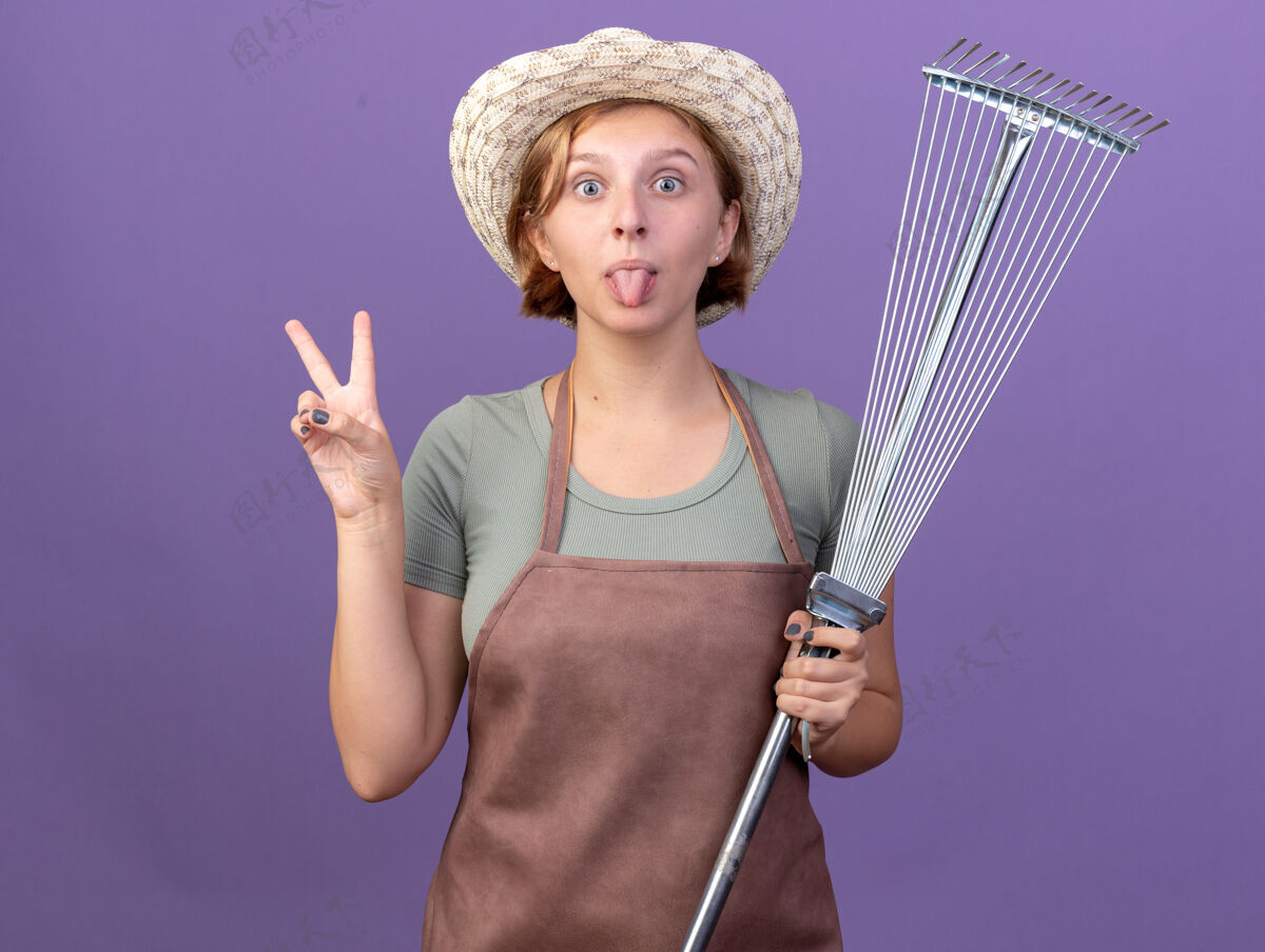 卡住令人印象深刻的年轻斯拉夫女园丁戴着园艺帽 伸出舌头 拿着树叶耙 在紫色上做着胜利的手势斯拉夫印象胜利
