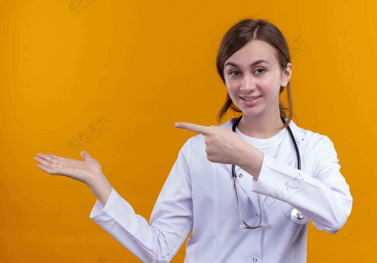 长袍高兴的年轻女医生穿着医用长袍和听诊器指着左边 在孤立的橙色空间显示空手橙色壁板显示