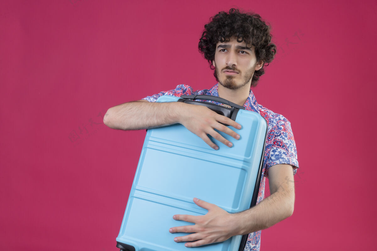 粉色严肃的年轻英俊的卷发旅行者男子举行孤立的粉红色空间与复制空间手提箱太空复制男人