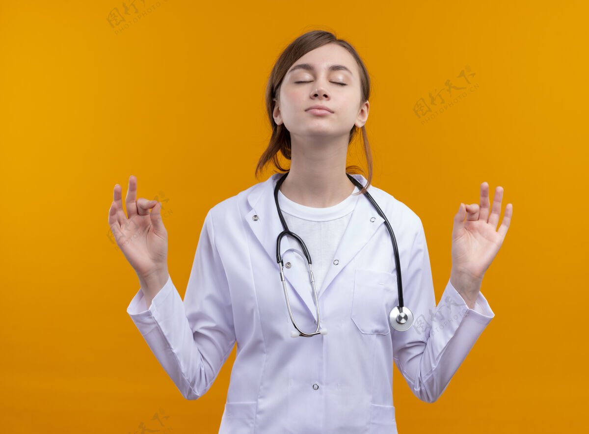 医生安静的年轻女医生 穿着医用长袍 戴着听诊器 闭着眼睛在孤立的橙色空间做着“ok”的手势可以女性听诊器