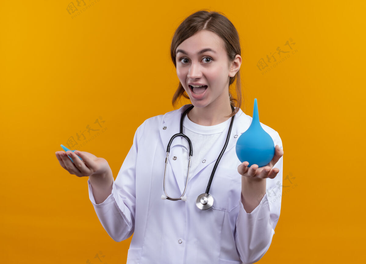 听诊器快乐的年轻女医生穿着医用长袍 手持听诊器在孤立的橙色空间灌肠长袍医疗女性