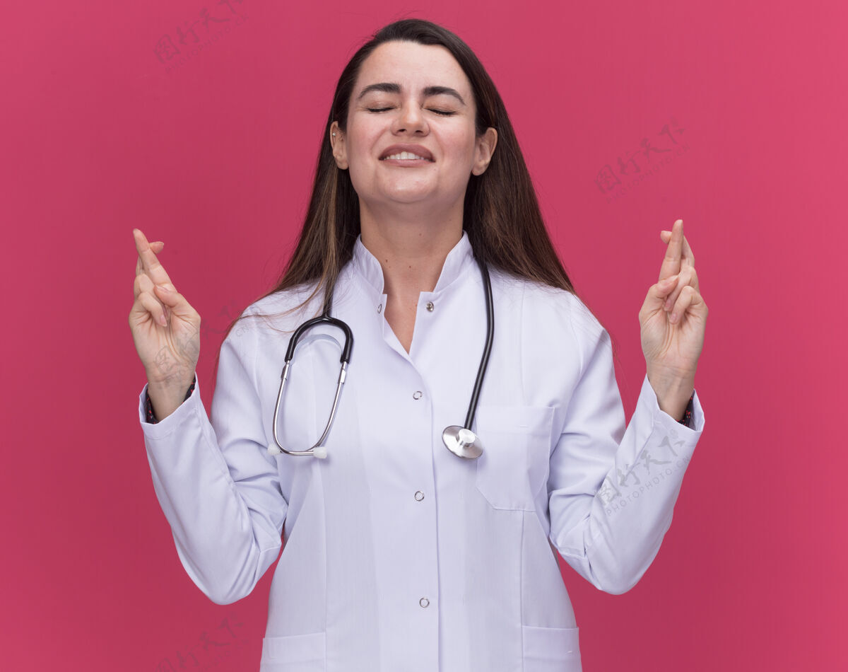 年轻焦急的年轻女医生穿着带听诊器的医用长袍 交叉着手指站在粉红色的床上长袍听诊器交叉