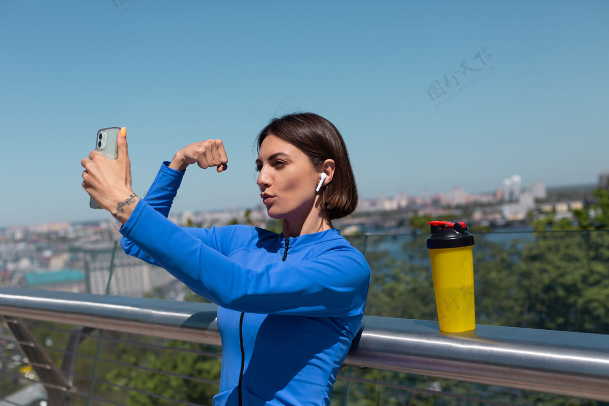 体育穿着蓝色运动服的年轻女子在炎热阳光明媚的早晨 戴着无线耳机和手机 在桥上为社交活动拍摄自拍视频 展示她的肌肉和肱二头肌健身自我运动员