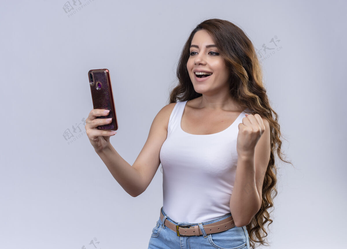 女孩快乐的年轻漂亮女孩拿着手机看着它 举起拳头手机年轻握着