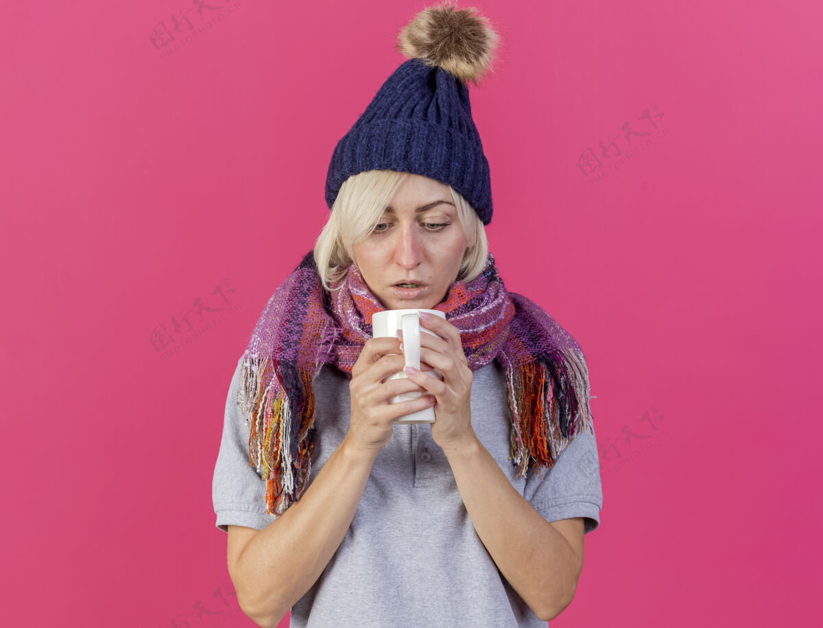 穿震惊的年轻金发病斯拉夫妇女戴着冬季帽子和围巾举行 并期待着在粉红色墙上的复制空间孤立的杯子年轻人帽子围巾