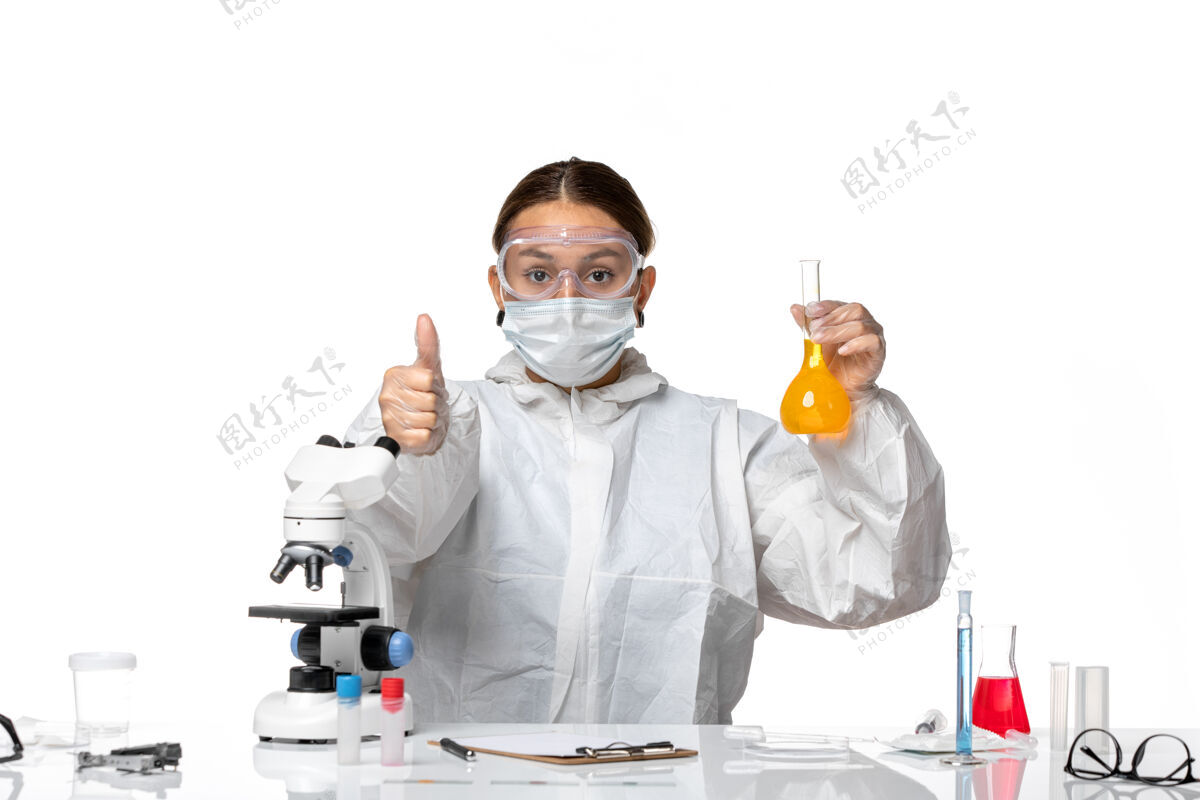 白色正面图：女医生 穿着特制的工作服 戴着口罩 拿着装有黄色溶液的烧瓶 背景是浅白色的冠状病毒大流行病毒大拇指朝上套装医生