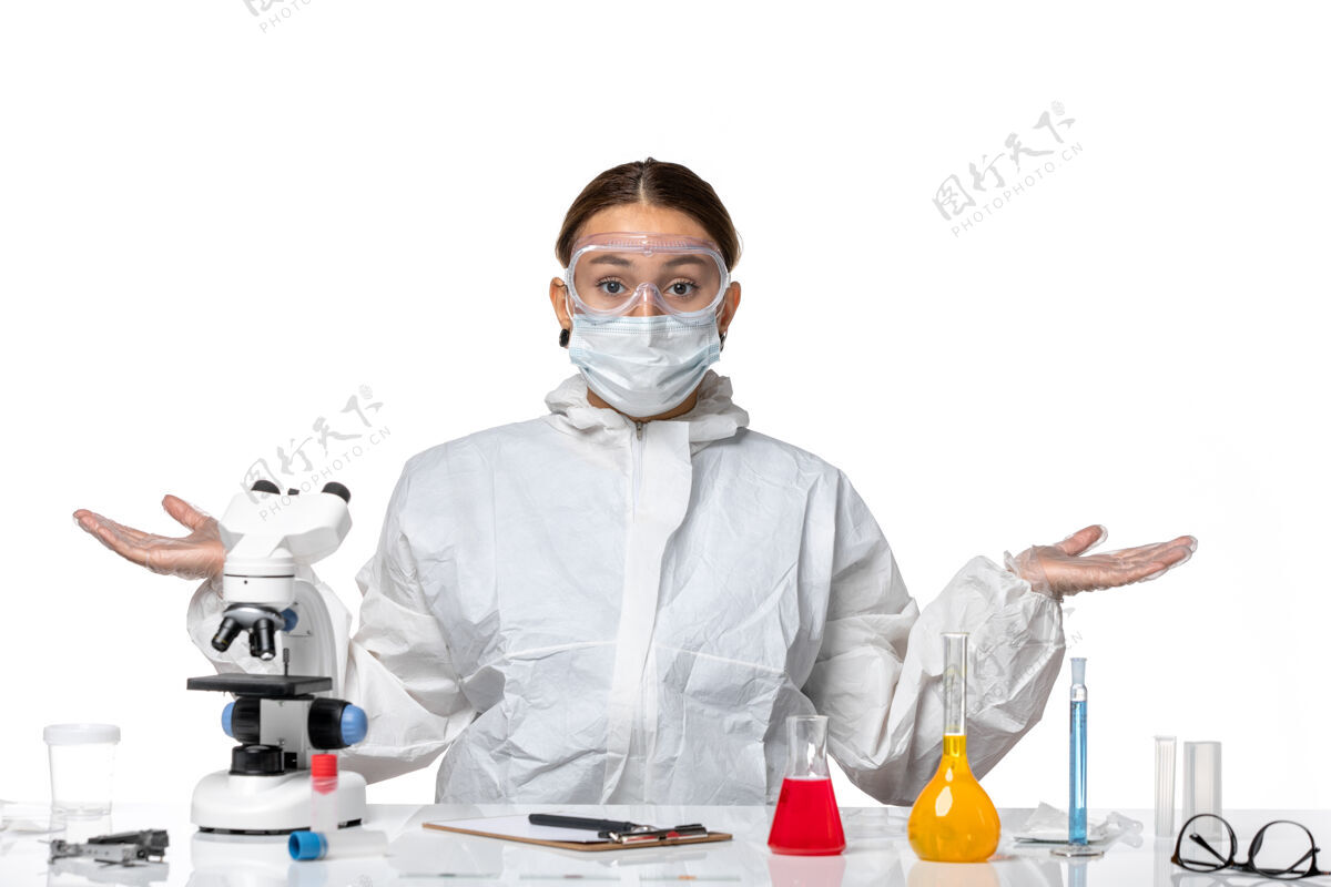 耸肩正面图：穿着防护服 戴着口罩的女医生坐在白色办公桌上解决病毒-健康大流行医疗实验服视野