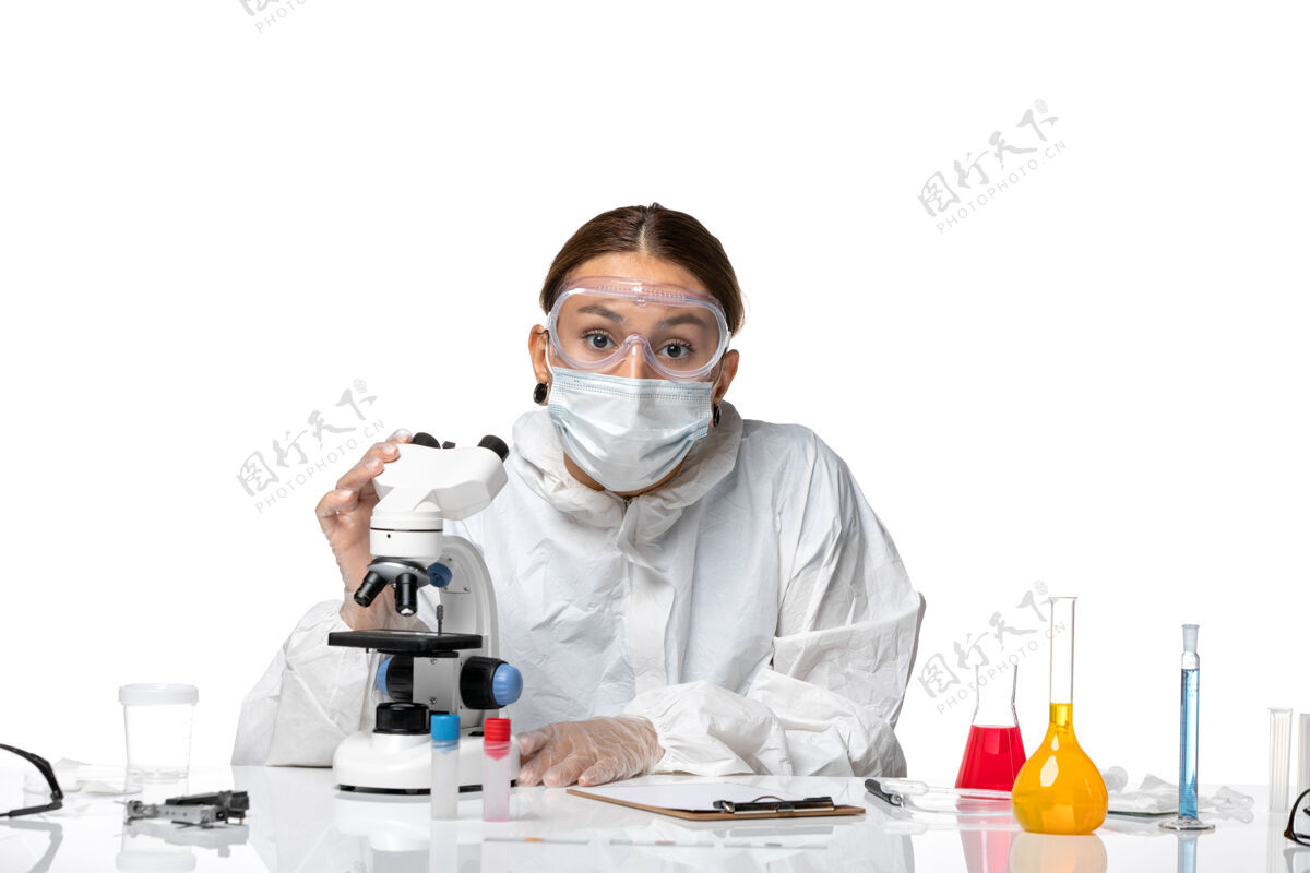 药品前视图：女医生穿着防护服 戴着口罩 用显微镜观察一种白色背景的健康冠状病毒大流行专业实验室外套女医生