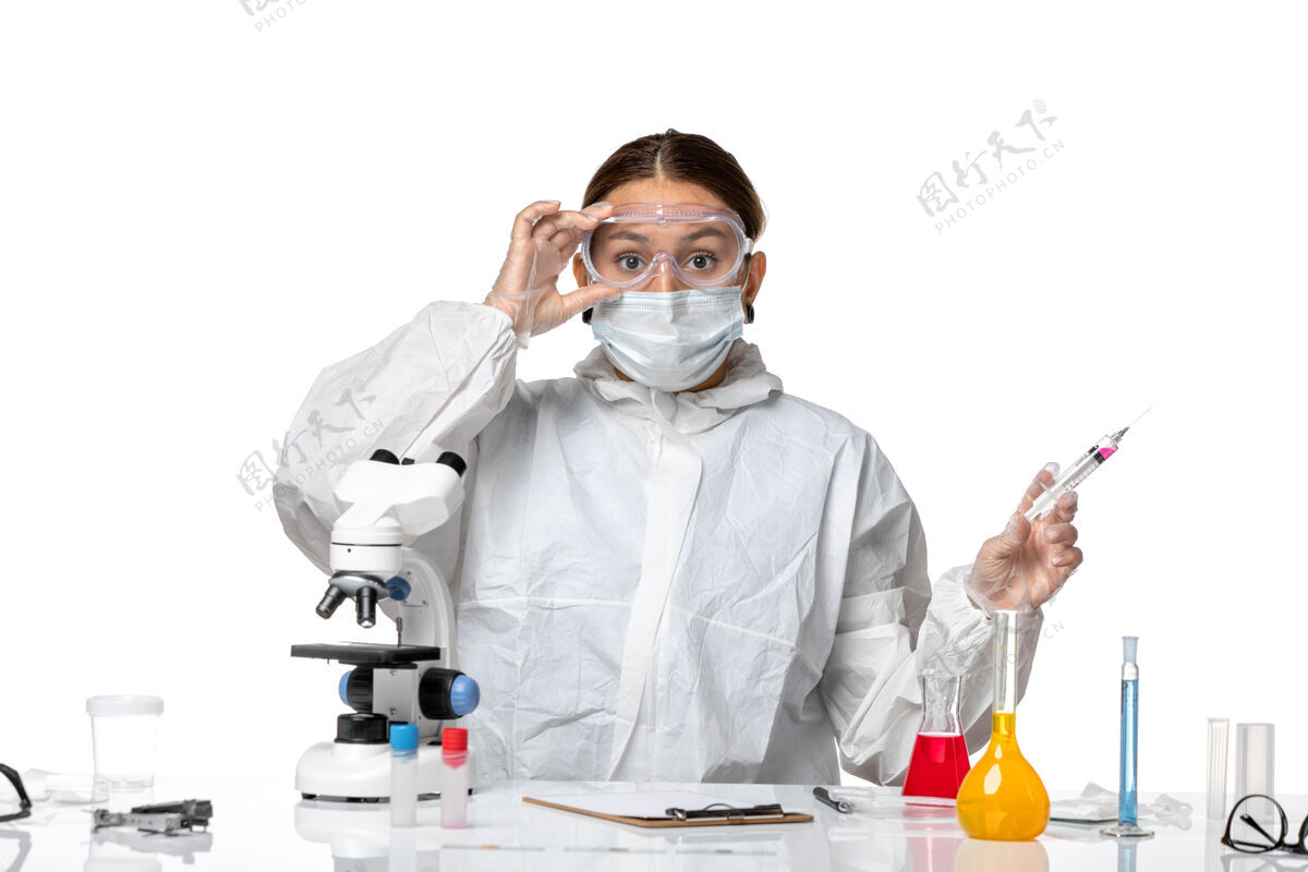 面罩正面图：穿着防护服 戴着口罩的女医生手持注射在浅白色背景的冠状病毒大流行病毒上人套装医生