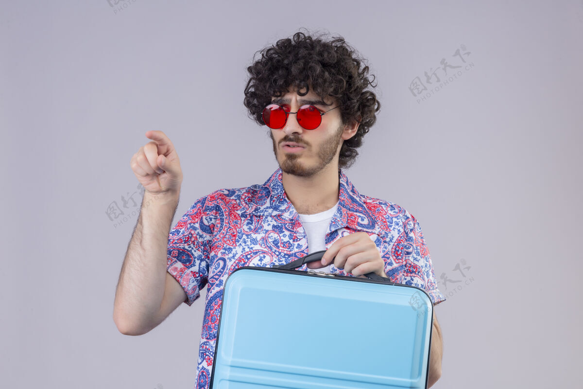 困惑困惑的年轻英俊的卷发旅行者戴着太阳镜拿着手提箱 指着前面有复印空间的孤立的白色空间卷发复制旅行者