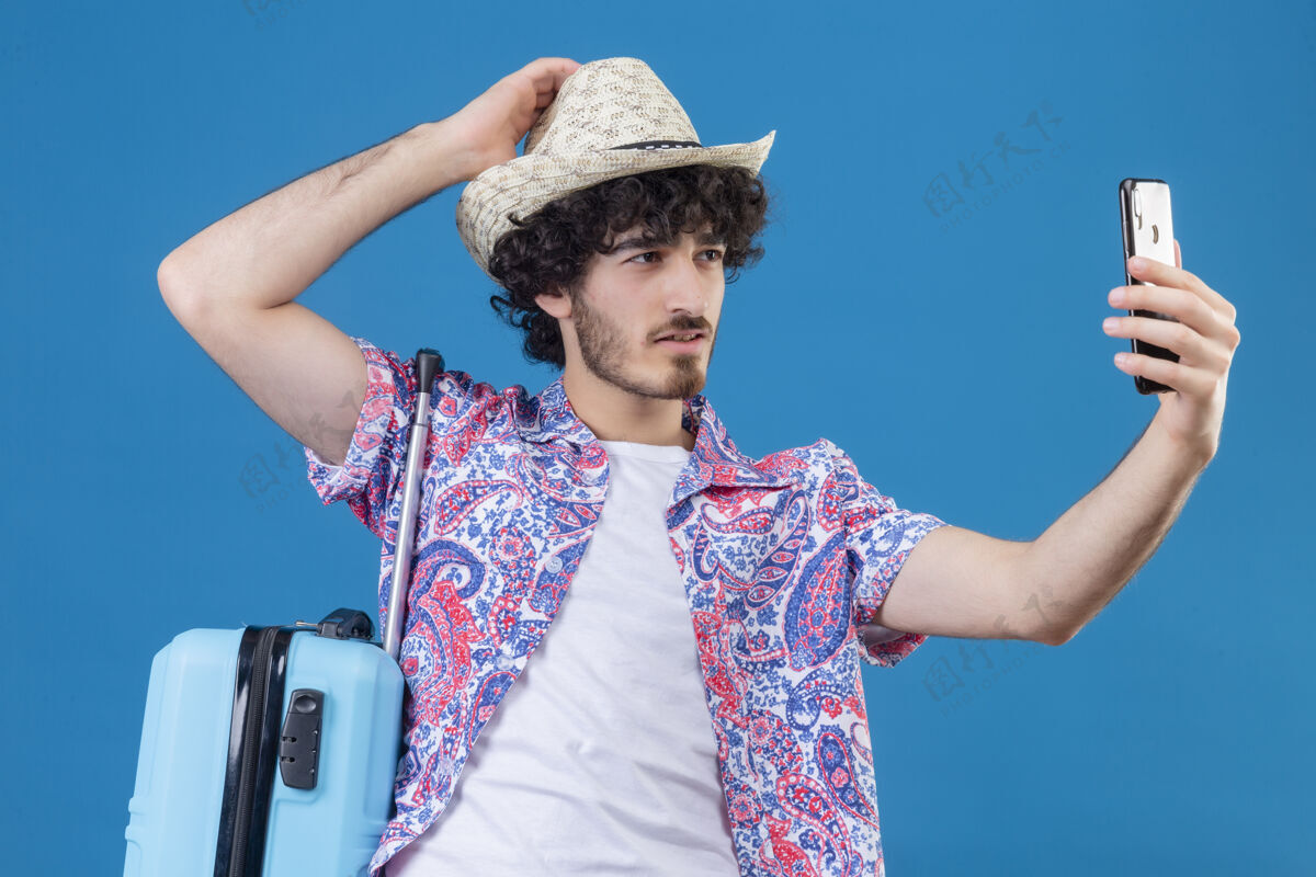 旅行者自信的年轻帅气卷发旅行者戴着帽子自拍手放在帽子上带着手提箱在与世隔绝的蓝色空间帅气手卷发