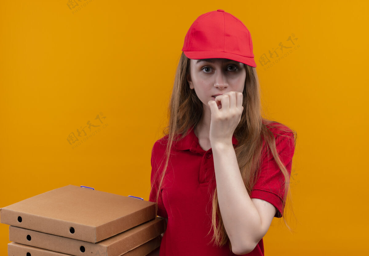 持有焦急的年轻送货女孩 穿着红色制服 手放在嘴唇上 手拿着包裹 站在孤立的橙色空间里橙色交货焦虑