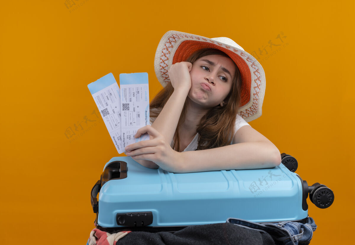 胳膊恼怒的年轻旅客女孩戴着帽子拿着机票 把手臂放在箱子上孤立的橙色空间手提箱橙色戴着