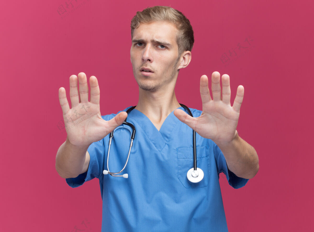 抱着穿着医生制服 手持听诊器的年轻男性医生 手伸向隔离在粉红色墙上的摄像机站着穿着制服