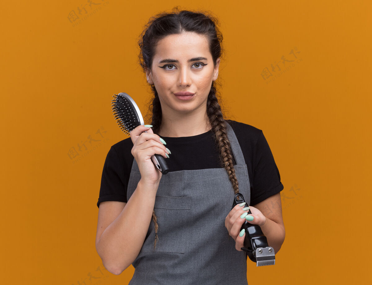 站着令人印象深刻的年轻女理发师穿着制服拿着梳子和剪子孤立在橙色墙上脸印象人