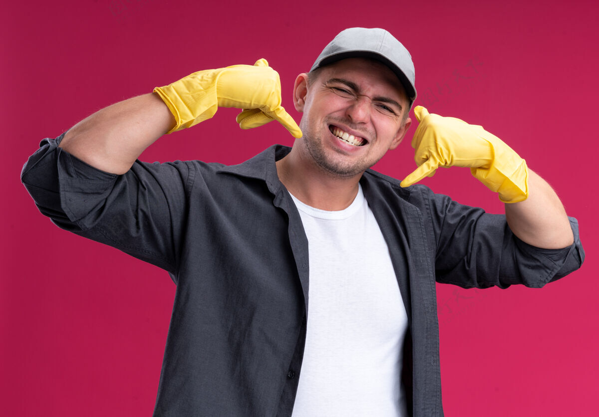 感觉微笑着的年轻帅气的清洁工 穿着t恤 戴着帽子 戴着手套 在粉红色的墙上显示着电话的手势穿着公民微笑