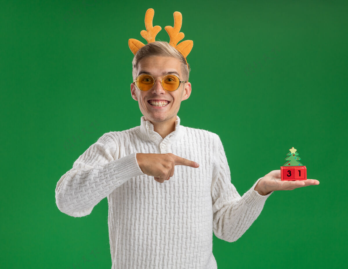 头带快乐的年轻帅哥戴着驯鹿鹿角头带戴着眼镜拿着并指着圣诞树玩具和日期看着隔离在绿色背景上的相机举行绿色鹿角