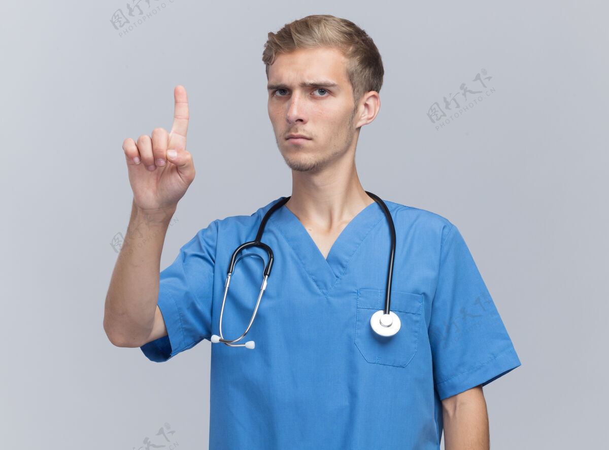 表情严格年轻的男医生穿着医生制服用听诊器指着隔离在白色墙上的上感情站人