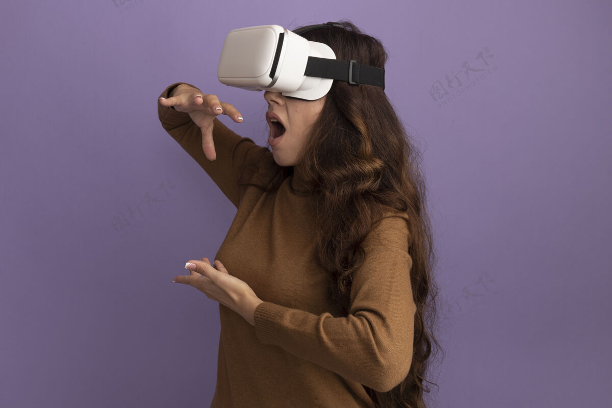姿势惊讶的年轻漂亮女孩戴着虚拟现实耳机假装拿着紫色墙上孤立的东西穿着脸市民
